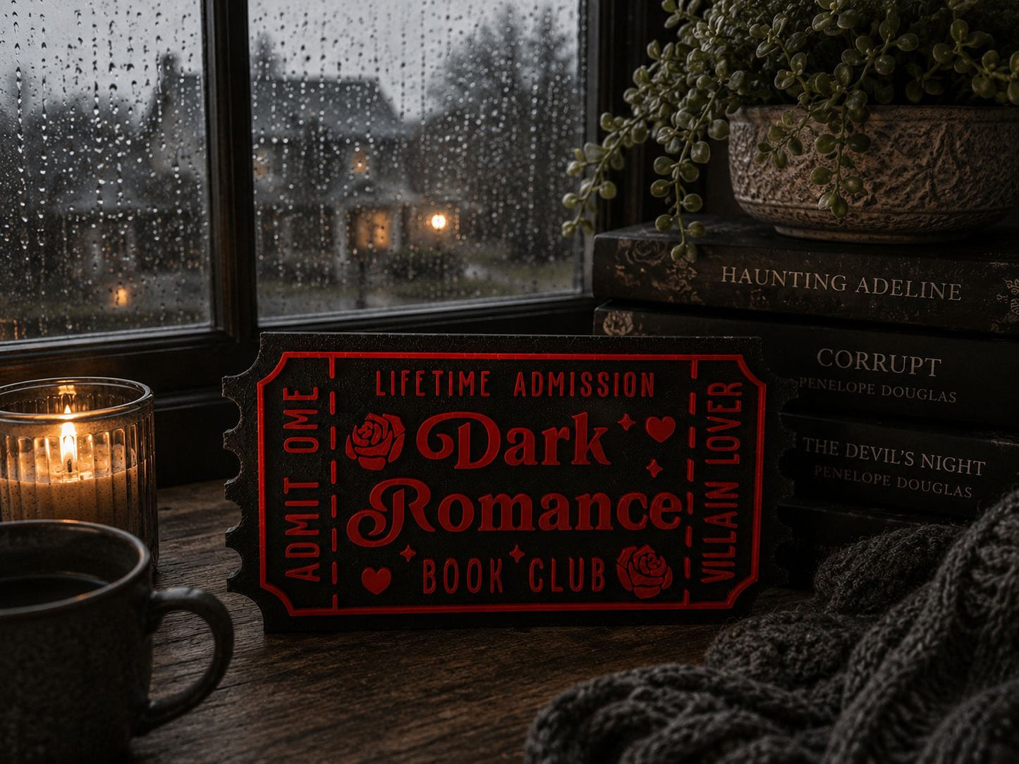 a cozy scene with a red ticket-shaped sign for "Dark Romance Book Club" placed on a wooden surface, surrounded by books, candles, and a potted plant.