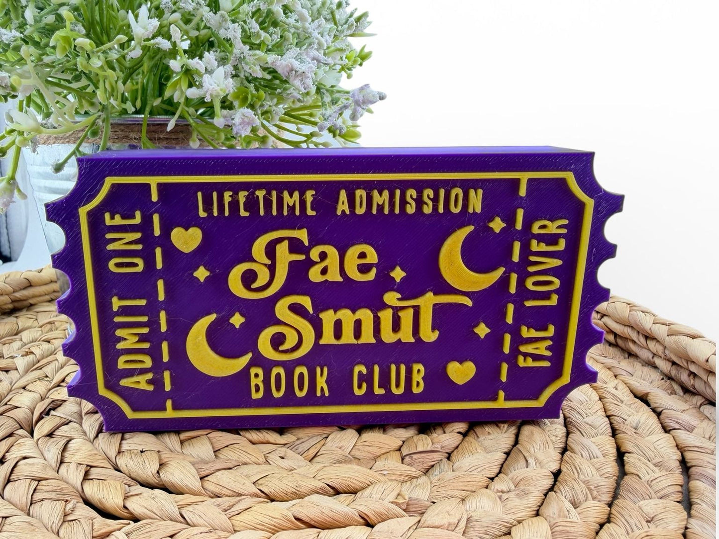 A purple ticket-shaped sign with gold lettering, displaying the text "Lifetime Admission Fae Smut Book Club" and two crescent moons, placed on a woven mat.