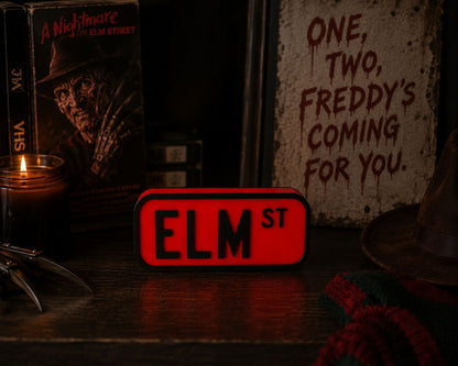 a collection of items related to the horror movie "A Nightmare on Elm Street", including a red sign with the word "ELM ST" on it, a book, a candle, and a hat.