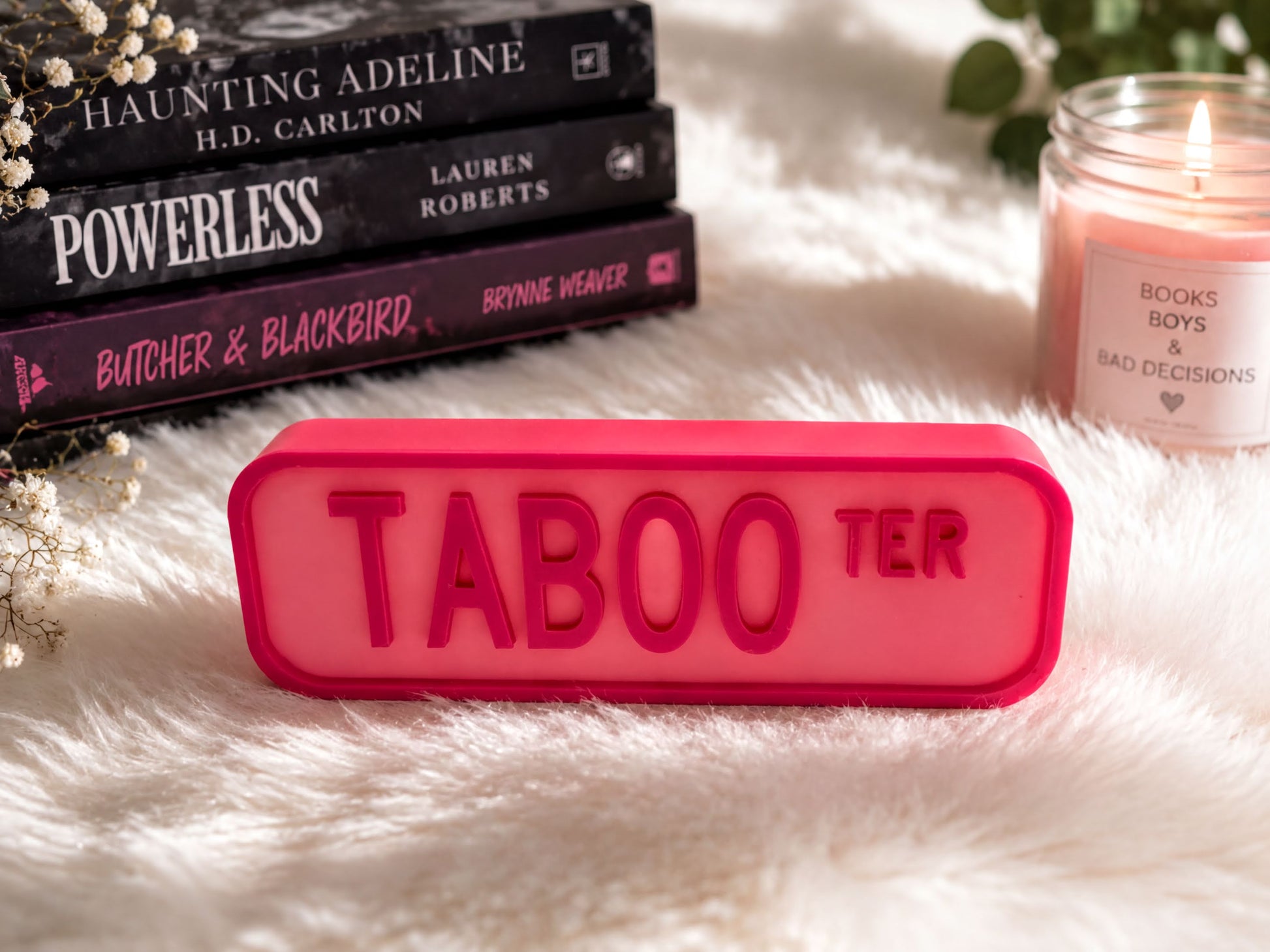 a pink "TABOO TER" sign in the foreground, surrounded by a stack of books, a candle, and some flowers on a white fur surface.