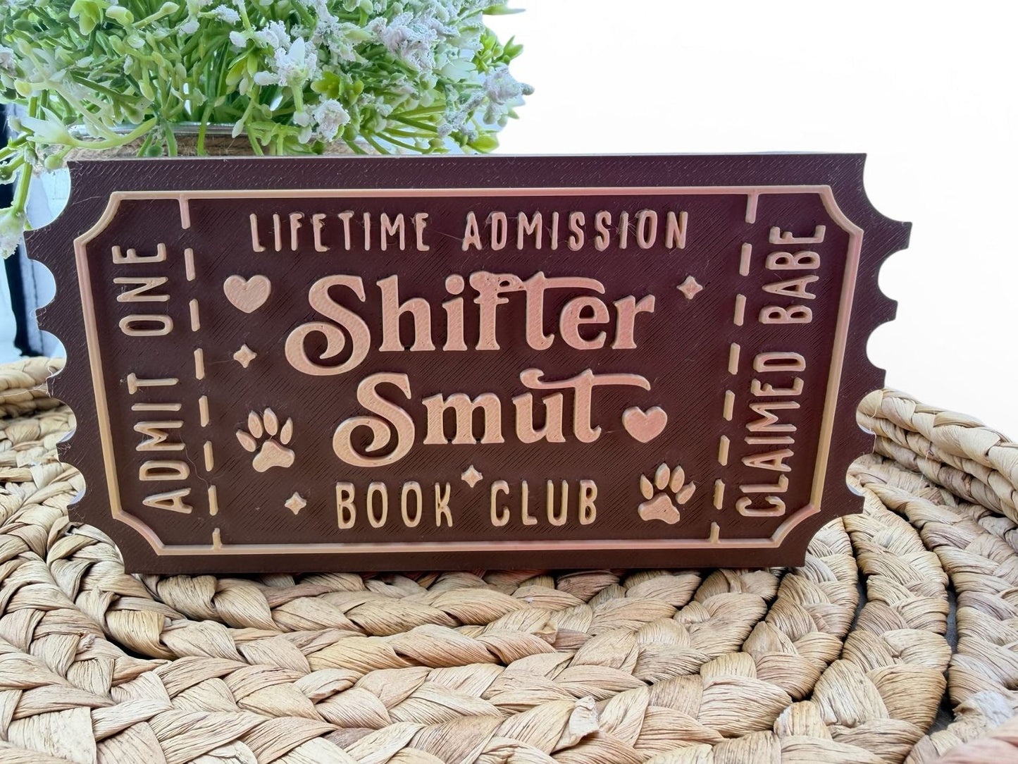 A brown wooden sign with the text "Shift Smut Book Club" on it, placed on a woven mat.