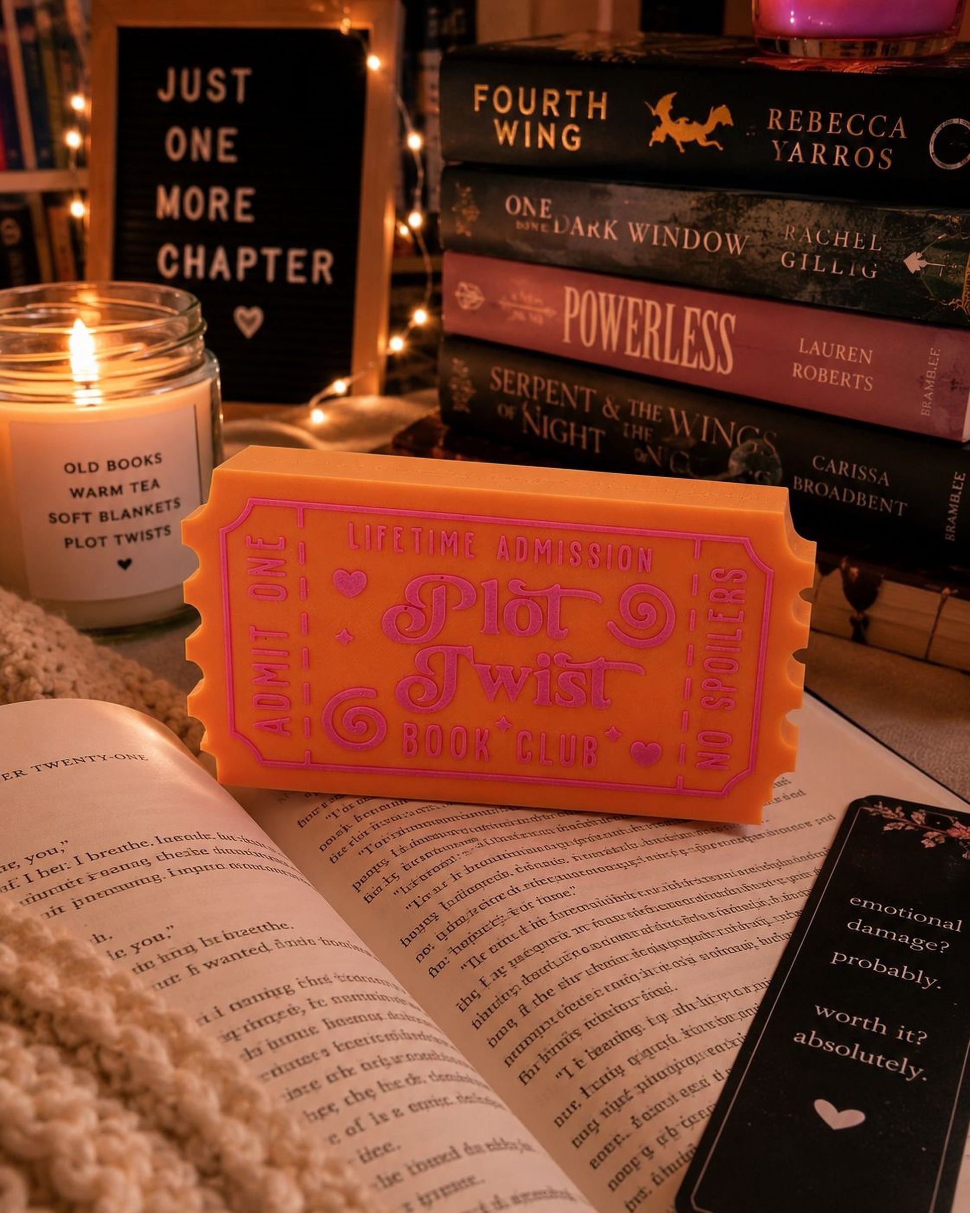 a cozy scene with a stack of books, a candle, and a pink ticket-shaped book club sign. The books are titled "Just One More Chapter" and "Powerless", while the candle appears to be a soft tea candle. The pink ticket-shaped book club sign reads "Lifetime Admission" and "No Spoilers". The open book in the foreground has text that reads "I am not a bridesmaid", suggesting a romantic or intimate atmosphere.