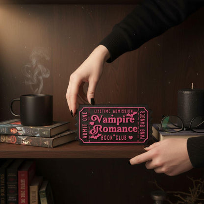 A hand is reaching for a book titled "Vampire Romance" on a shelf, surrounded by other books and a mug.