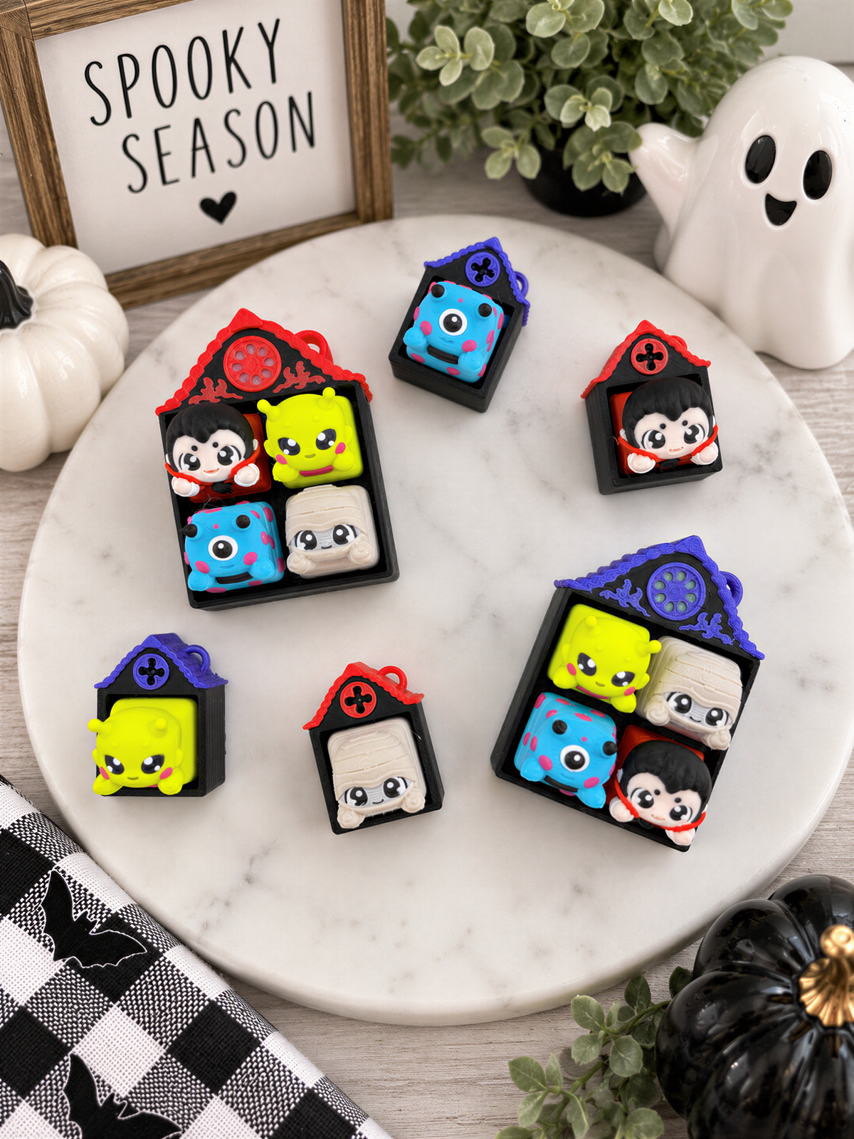 a collection of colorful, cartoonish keychain-like objects arranged on a white marble surface. There are several small houses, each with unique designs and decorations, including a ghost and a pumpkin. The objects are arranged in a playful and whimsical manner, creating a visually appealing display.