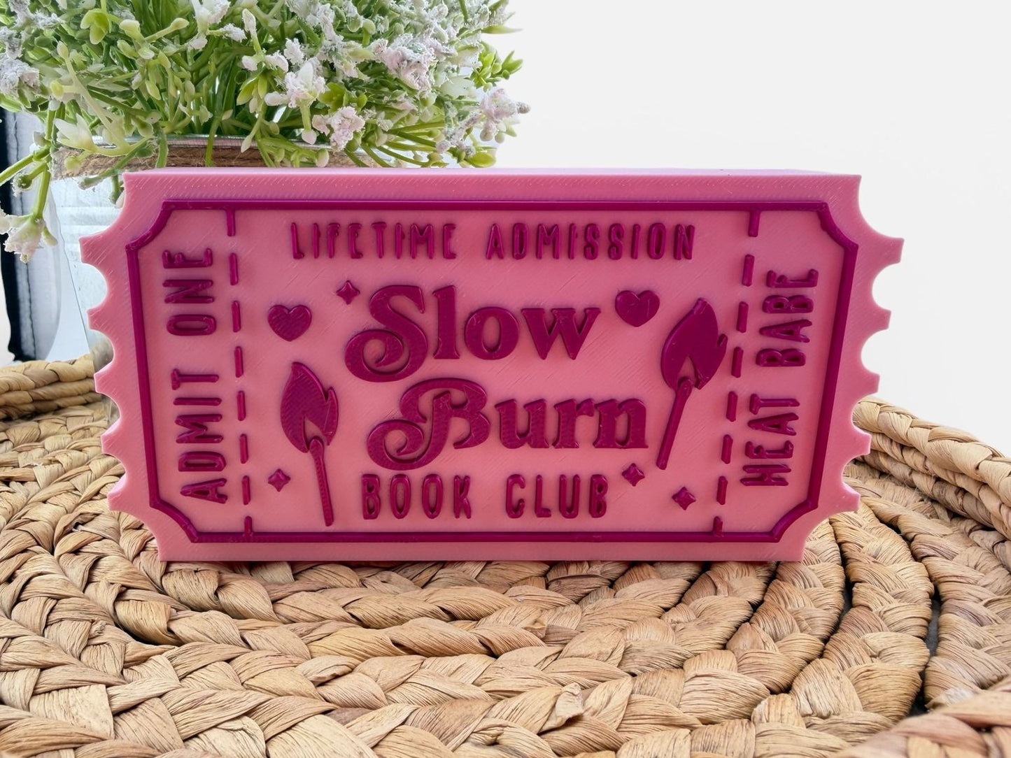 A pink ticket-shaped object with text that says "Lifetime Admission Slow Burn Book Club" on it, placed on a woven surface.