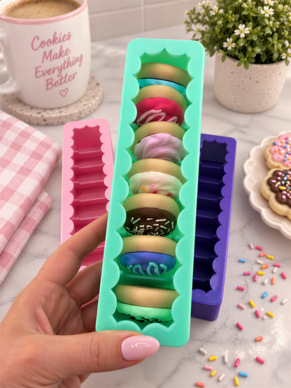 A hand holding a green container filled with colorful, scalloped-edge cookies, surrounded by other cookies, a mug, and a potted plant.