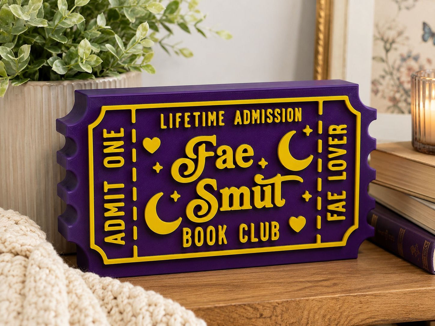 A purple and yellow ticket-shaped sign for the "Fae Smut Book Club" is displayed on a wooden surface, with a potted plant and books in the background.