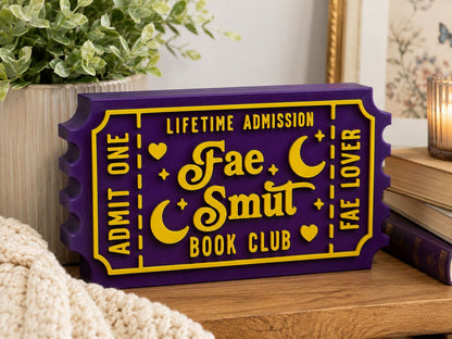 A purple and yellow ticket-shaped sign for the "Fae Smut Book Club" is displayed on a wooden surface, with a potted plant and books in the background.