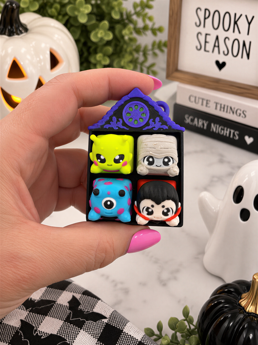 A hand holding a small toy with four different colored characters on it, surrounded by Halloween decorations and a framed sign.
