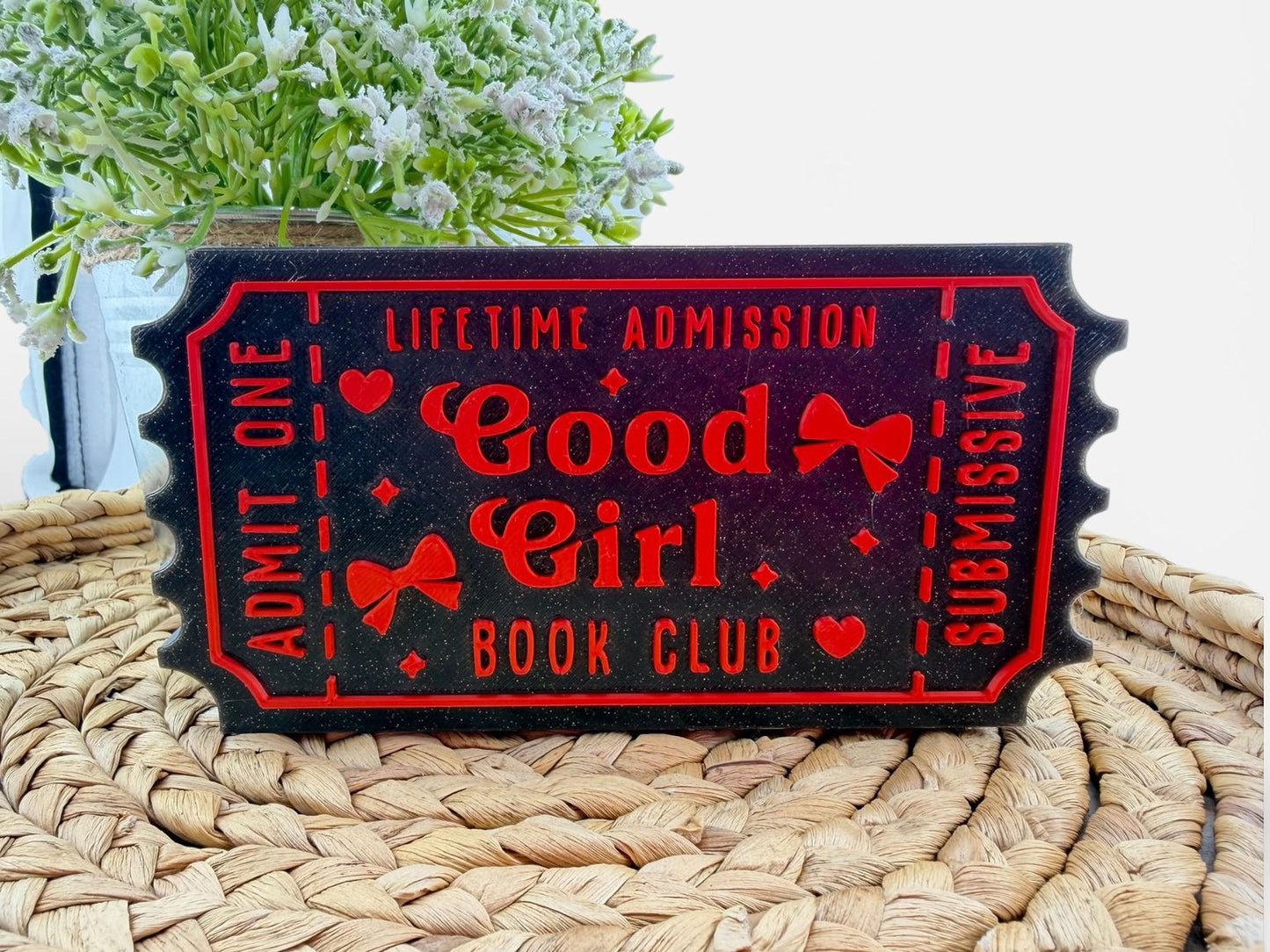 A black and red ticket-shaped sign with the text "Good Girl Book Club" on it, placed on a woven mat.