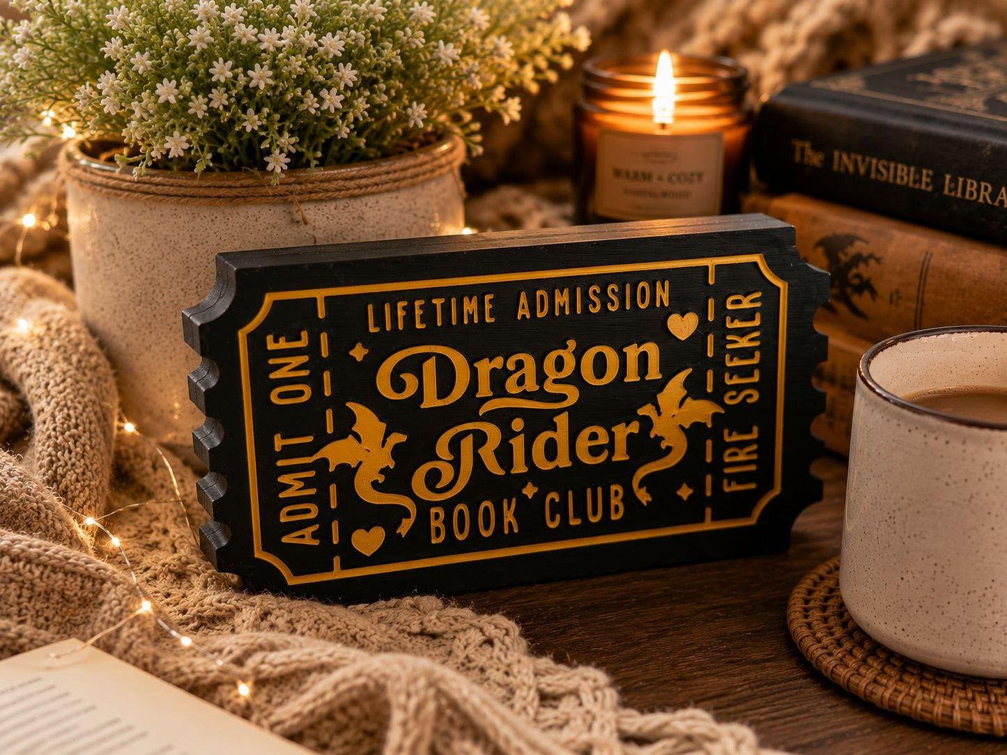 a black and gold ticket-shaped sign with the text "Dragon Rider Book Club" on it, surrounded by various items such as a candle, a mug, and a book.