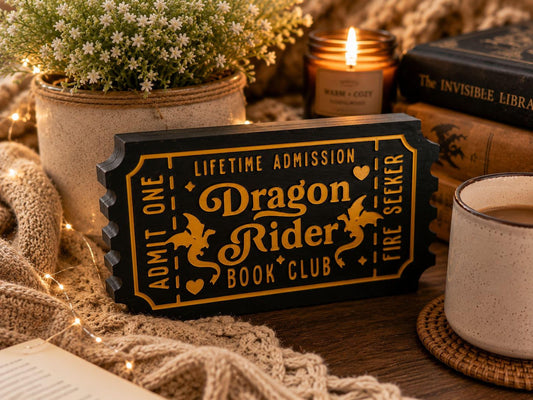 a black and gold ticket-shaped sign with the text "Dragon Rider Book Club" on it, surrounded by various items such as a candle, a mug, and a book.