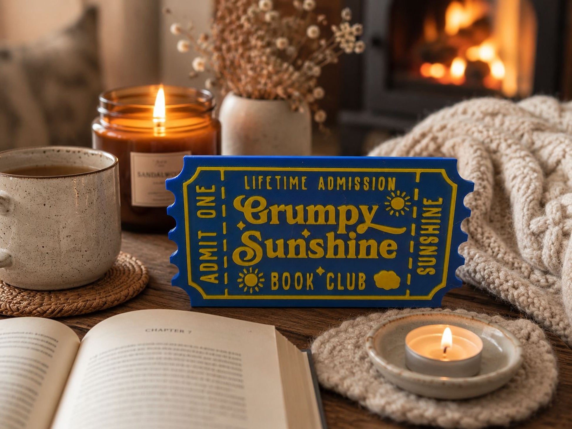 a cozy scene with a blue ticket-shaped sign that reads "Lifetime Admission Crumpy Sunshine Book Club", a lit candle, a mug, a book, and a vase with flowers.