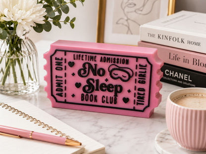 a pink ticket-shaped object with the text "No Sleep Book Club" on it, placed on a marble surface next to a notebook, pen, and a cup of coffee.