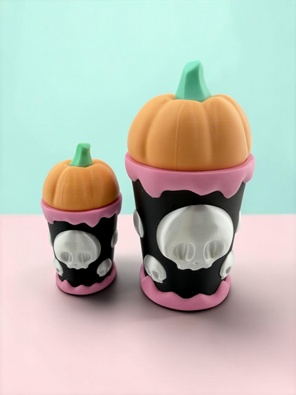 Two small, colorful pumpkin-shaped containers with skull designs on them, placed on a pink surface against a light blue background.