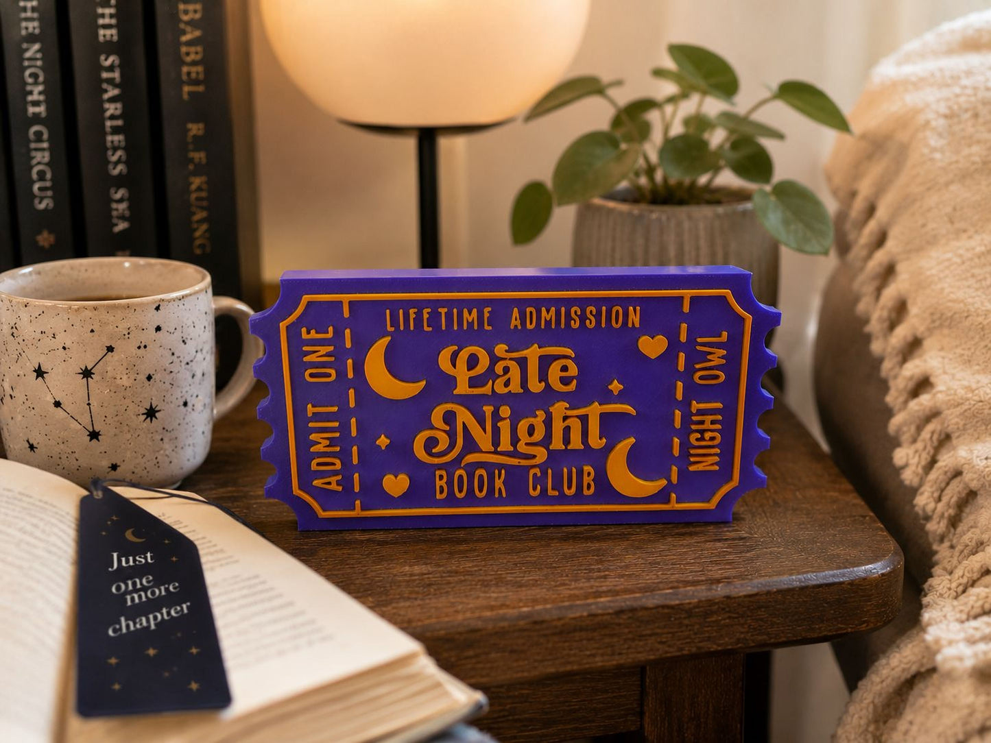 a purple ticket-shaped sign that says "Lifetime Admission Late Night Book Club" on a wooden table, with a mug, a lamp, and a plant nearby.