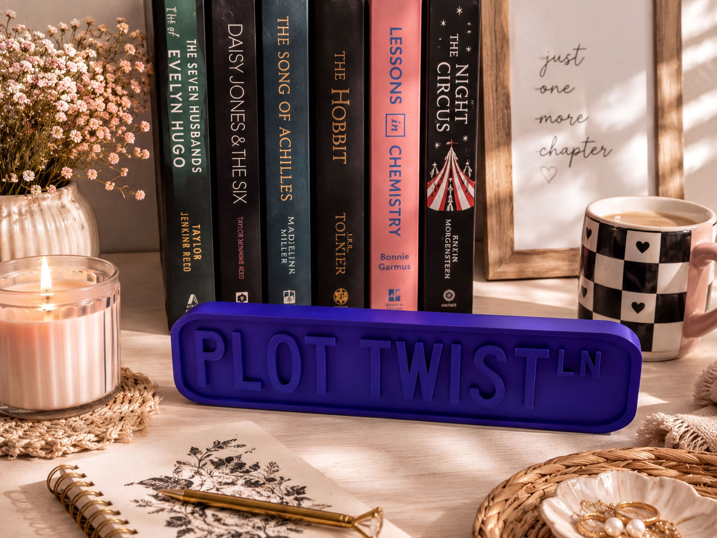 a collection of books, a candle, a mug, and a notebook on a desk, with a sign that says "PLOT TWIST LN".