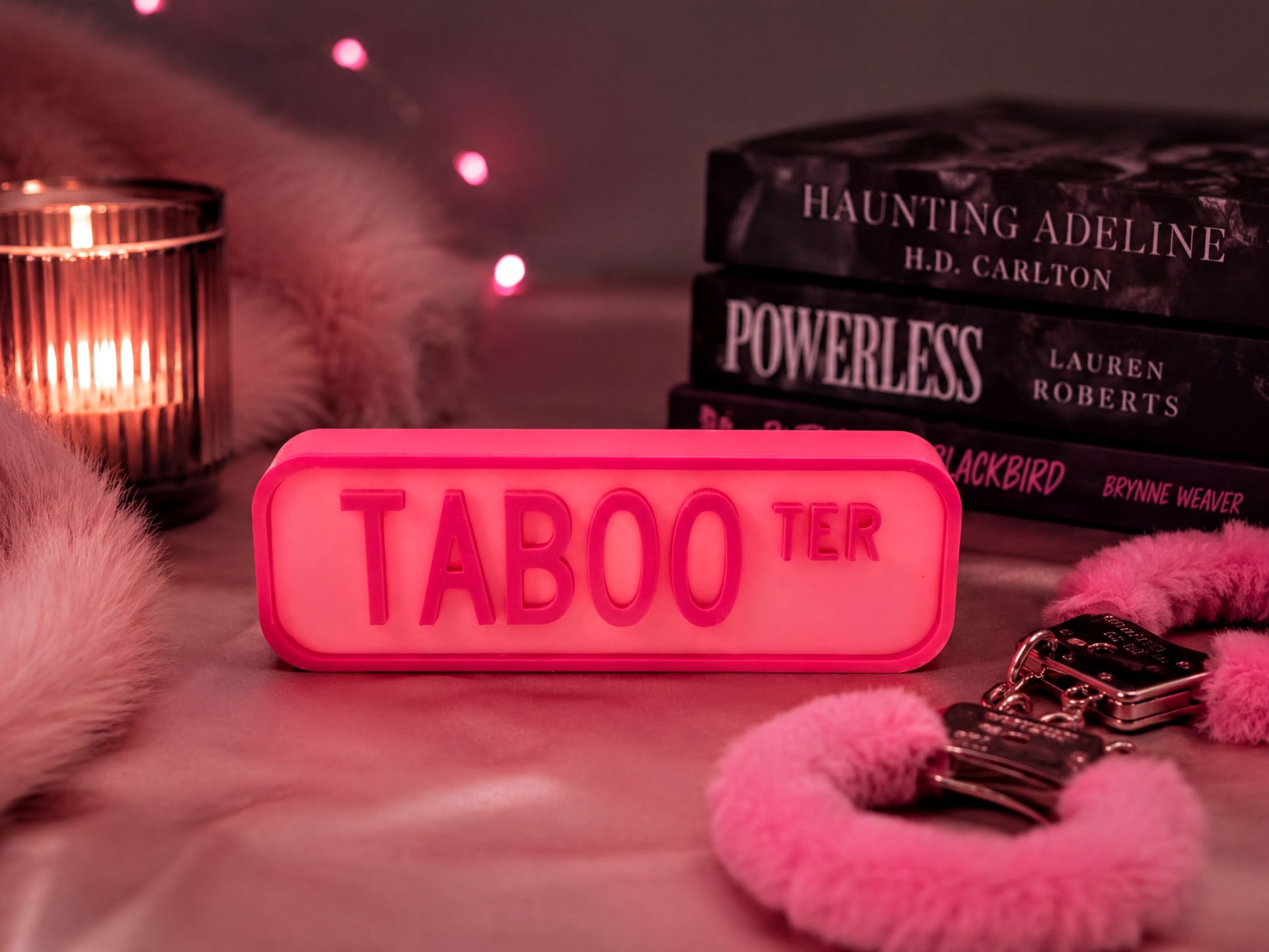 a pink "Taboo" sign, a stack of books, a lit candle, and some pink fur accessories, all arranged on a pink surface.