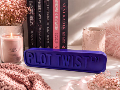 a collection of books, a candle, and a decorative object with the text "PLOT TWIST" on it, all arranged on a surface with pink flowers.