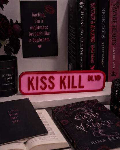 a shelf with various books, a pink sign that says "KISS KILL BLVD", and some decorative items such as a candle and a vase.