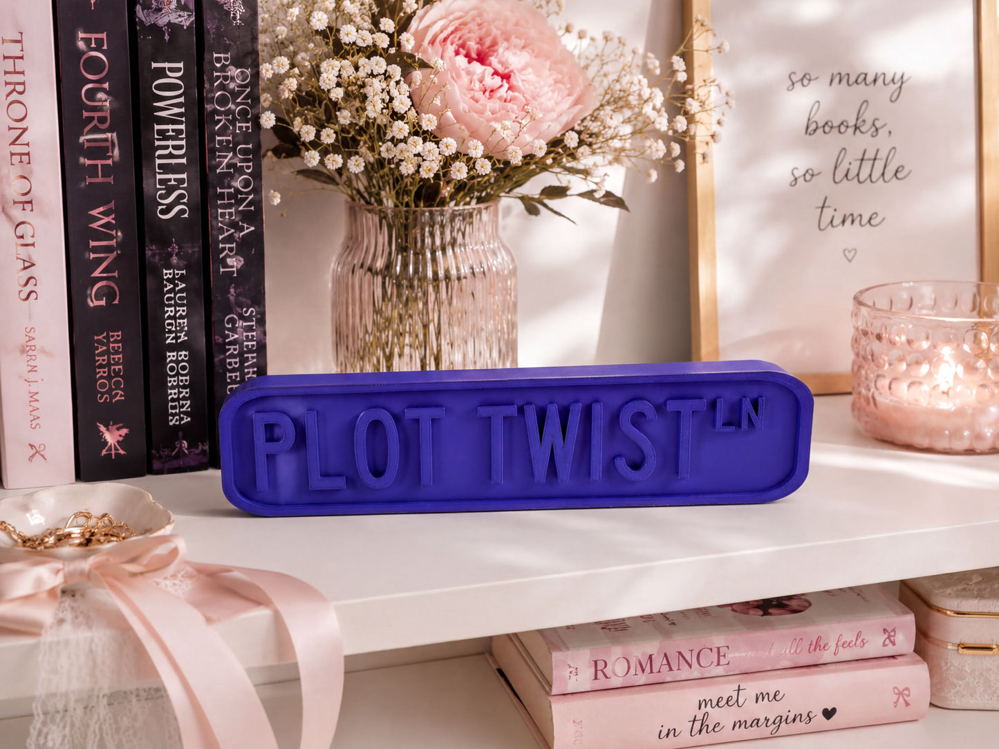 a shelf with various items, including a vase of flowers, books, and a purple sign that says "PLOT TWIST".