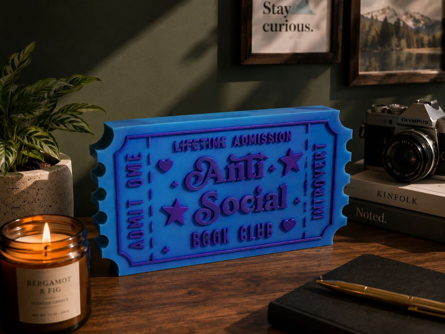 a blue ticket-shaped object with the text "Lifetime Admission Anti Social Book Club" on it, placed on a wooden desk. There is also a candle, a camera, and some books in the background.