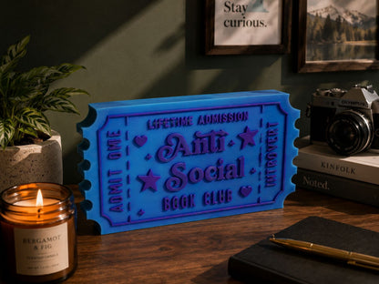a blue ticket-shaped object with the text "Lifetime Admission Anti Social Book Club" on it, placed on a wooden desk. There is also a candle, a camera, and some books in the background.