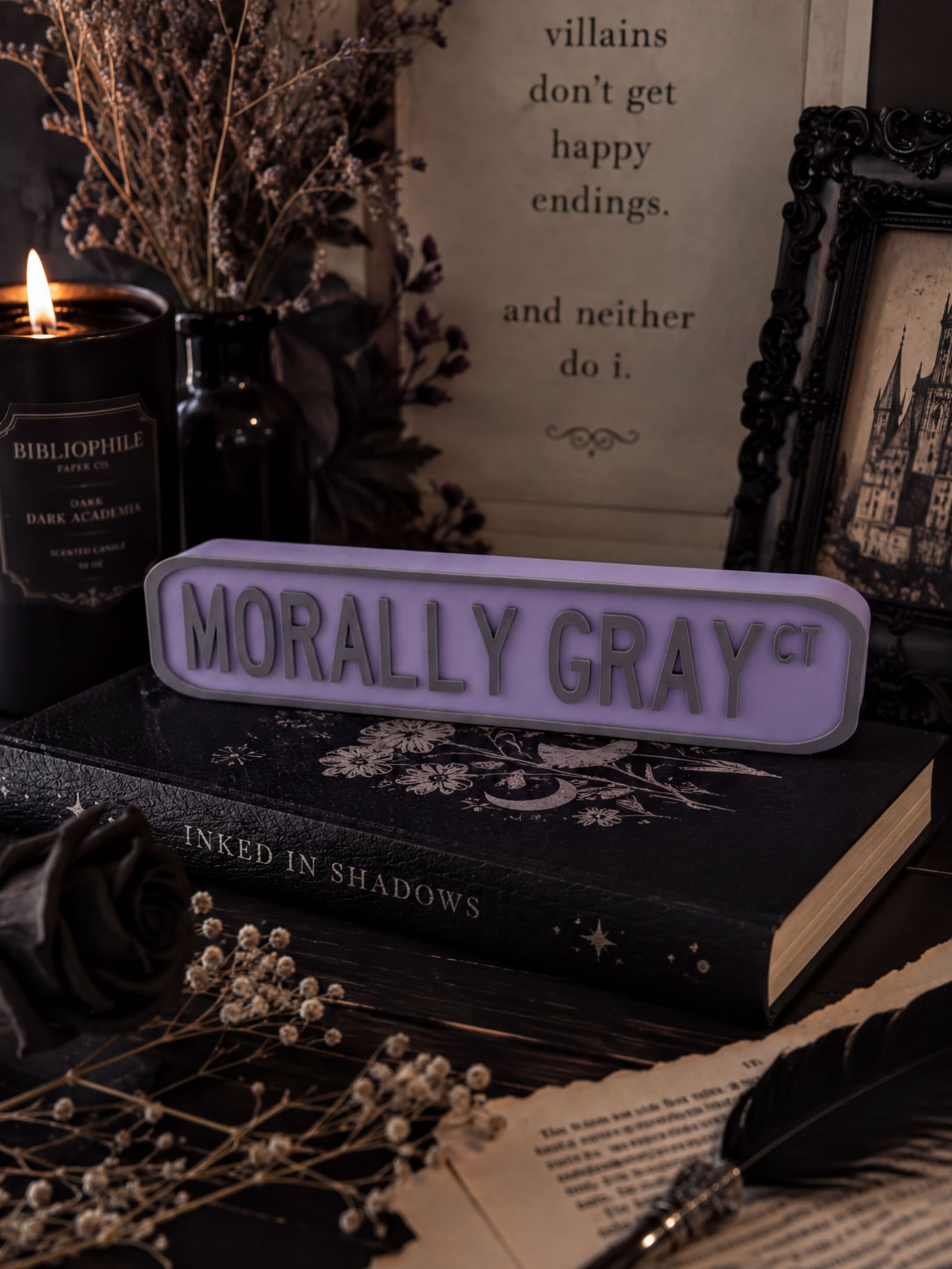 a dark and moody scene with a purple sign that reads "Morally Gray" placed on top of a black book. The background includes a candle, a vase with dried flowers, and a framed quote.