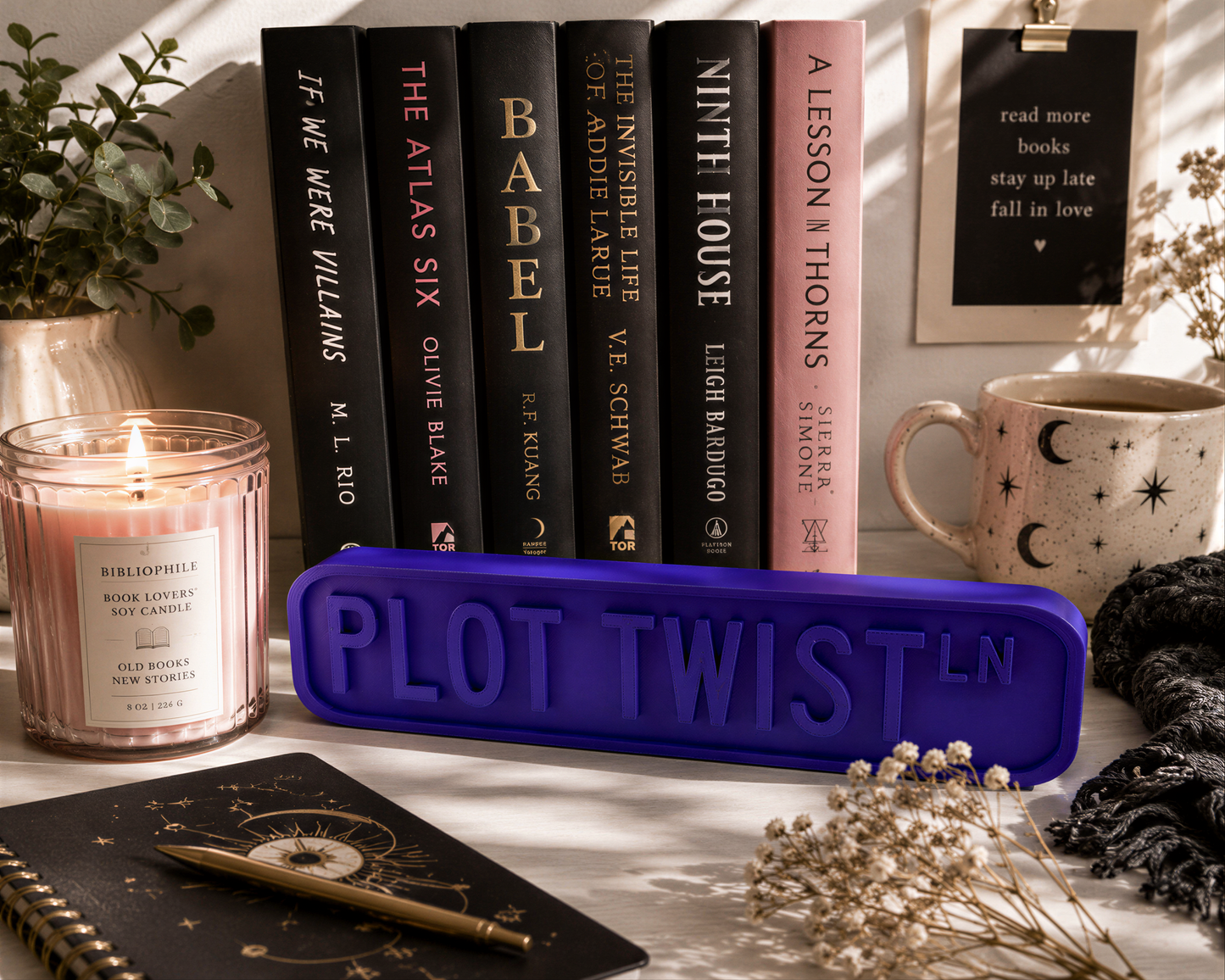 a collection of books, a candle, a mug, and a notebook arranged on a desk, with a purple sign that says "PLOT TWIST LN" in the foreground.