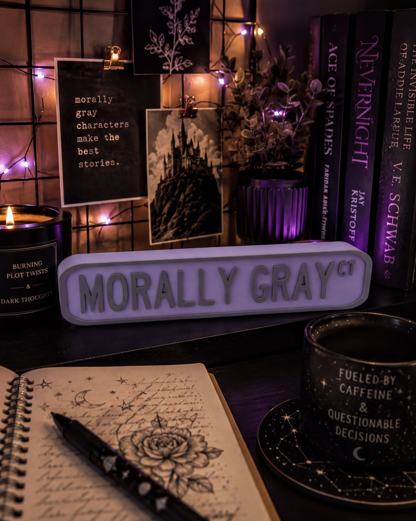 a desk with various items, including a notebook, a candle, a mug, and a sign that says "Morally Gray". There are also books and a plant on the desk.