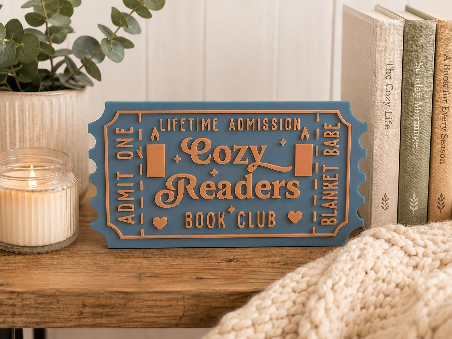 A blue and gold ticket-shaped sign for a cozy readers book club, placed on a wooden surface next to a candle and some books.
