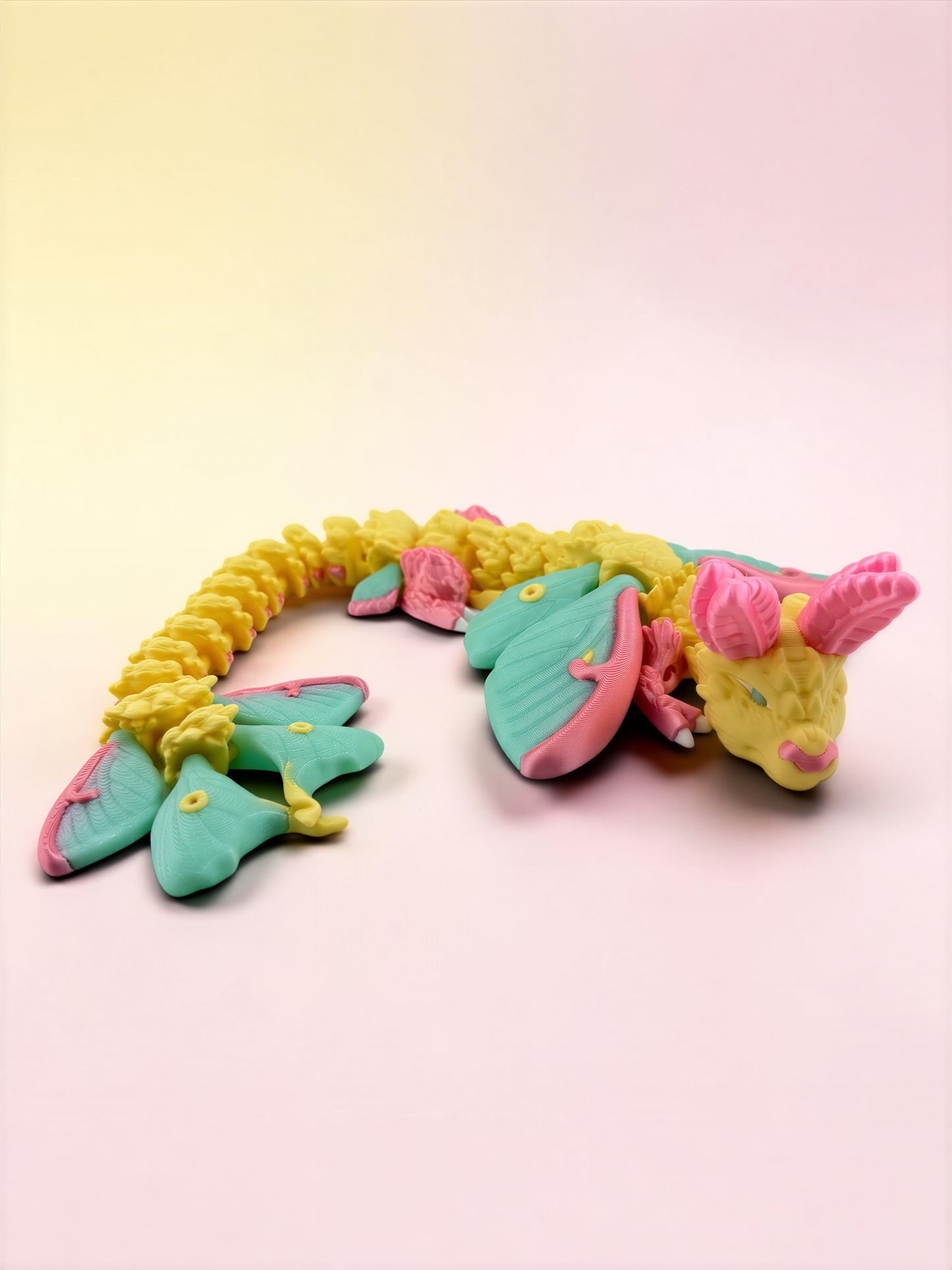 A colorful toy dragon made of plastic or clay, with a yellow body, green wings, and pink accents, is lying on a pink background.