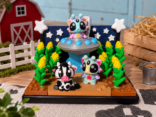 A whimsical display featuring three colorful toy figurines, including a cow, a cat, and a spaceship, set against a backdrop of a red barn, yellow flowers, and a starry night sky.
