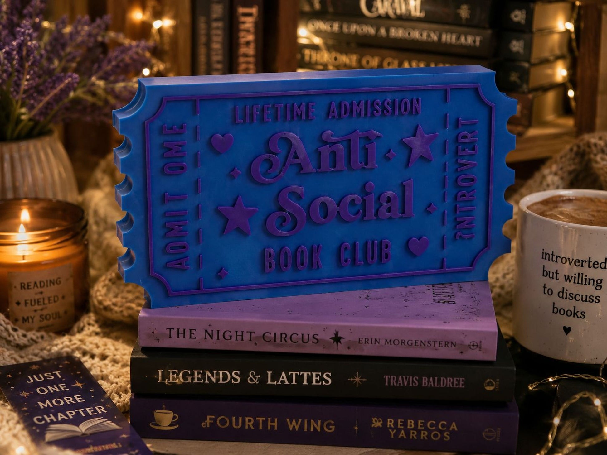 a blue ticket-shaped object with the words "Lifetime Admission Anti Social Book Club" written on it, surrounded by various books and a candle.
