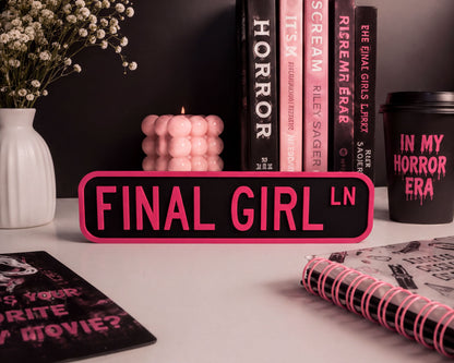 a desk with various items, including a pink sign that says "FINAL GIRL", a candle, a notebook, and a coffee cup.