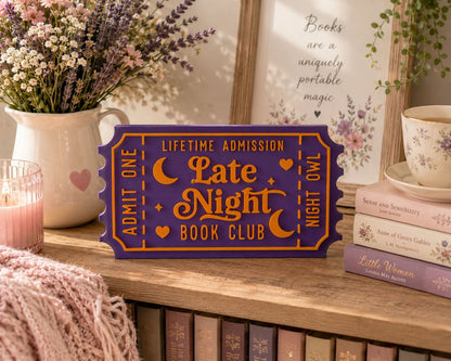 a purple ticket-shaped sign that reads "Lifetime Admission Late Night Book Club" on a wooden shelf, surrounded by books, a vase of flowers, and a framed quote.
