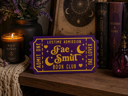 a purple ticket with gold lettering, placed on a wooden surface next to a candle, a vase of purple flowers, and a book.