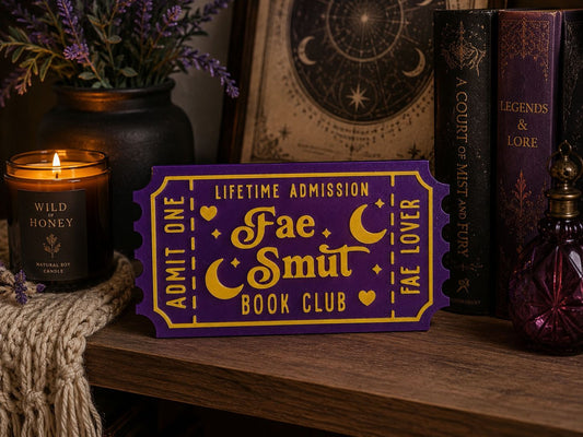 a purple ticket with gold lettering, placed on a wooden surface next to a candle, a vase of purple flowers, and a book.