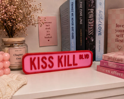 a shelf with various books, a pink sign that says "KISS KILL BLVD", and a vase with flowers.