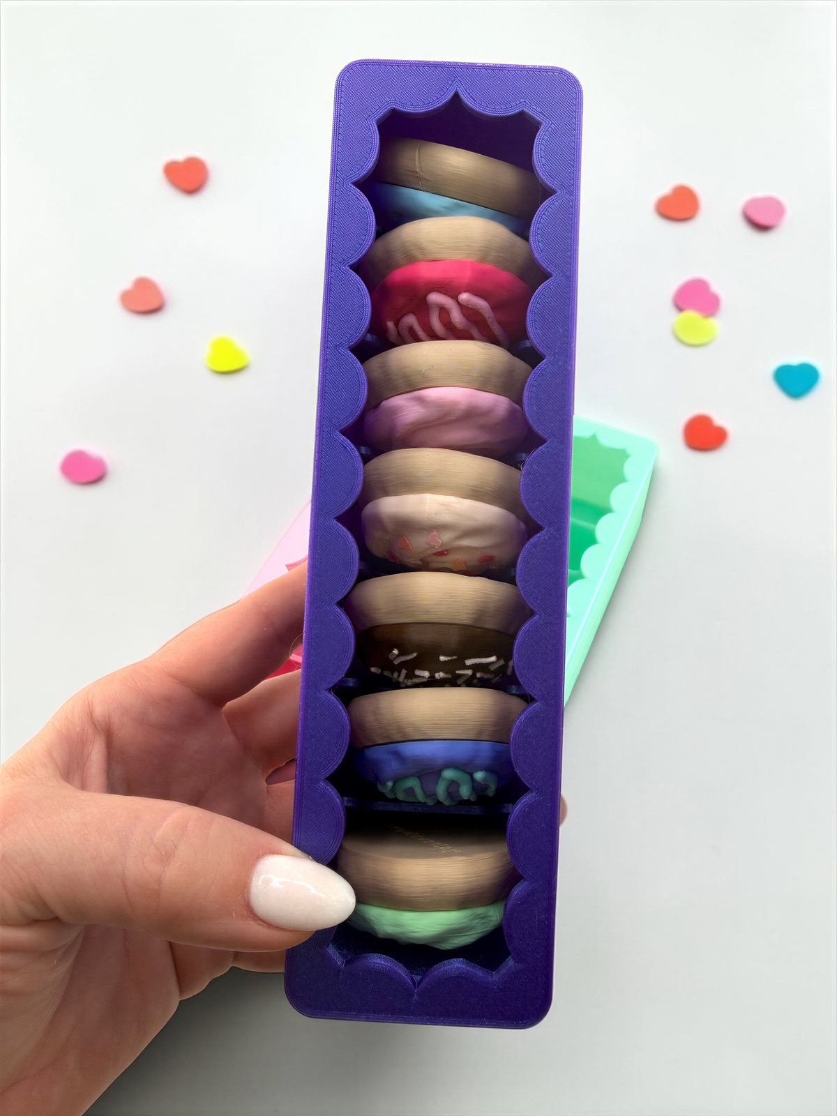 A hand holding a purple container filled with colorful heart-shaped cookies, surrounded by scattered heart-shaped cookies on a white background.