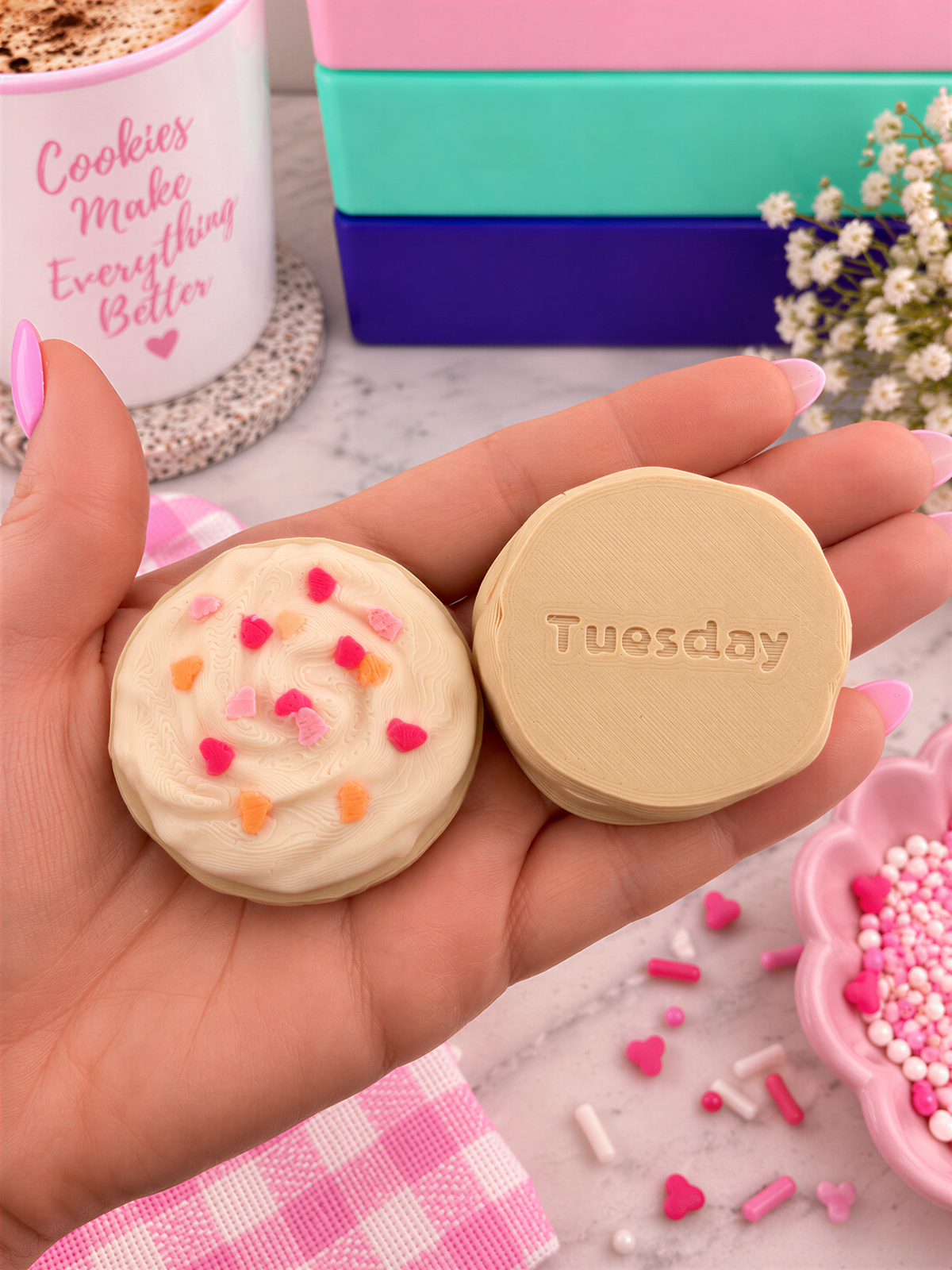 A hand holding two round cookies with sprinkles, one labeled "Tuesday". The cookies are placed on a table with a pink and white checkered napkin, a mug with the text "Cookies make everything better", and some pink and white sprinkles scattered around.