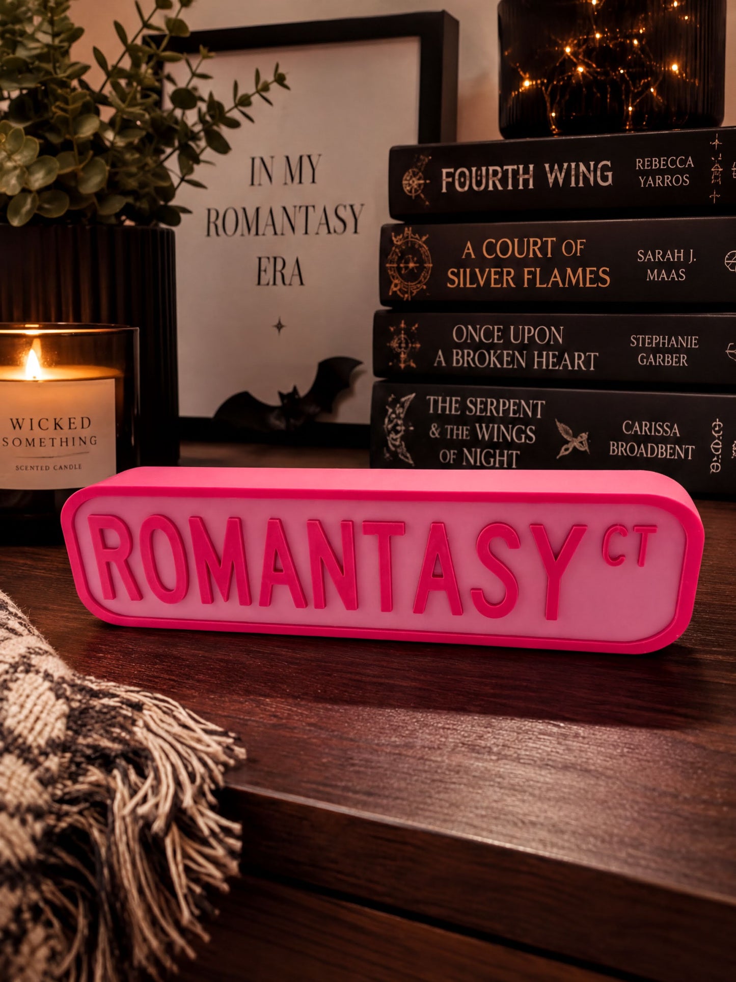 a pink sign with the word "ROMANTASY" on it, surrounded by various books and a candle on a wooden surface.
