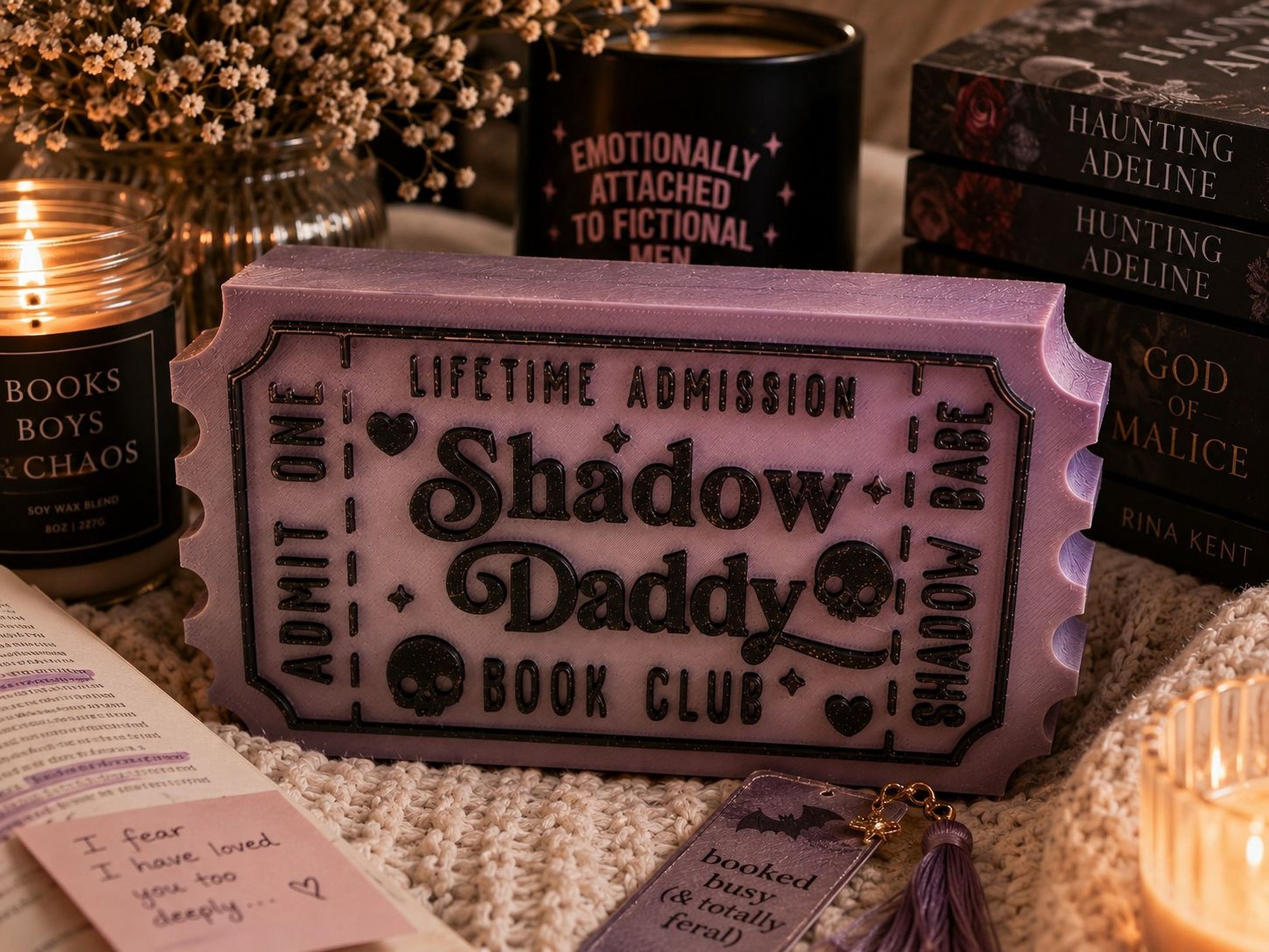a purple ticket-shaped object with the text "Lifetime Admission Shadow Daddy Book Club" on it, surrounded by various items such as candles, books, and a tassel.