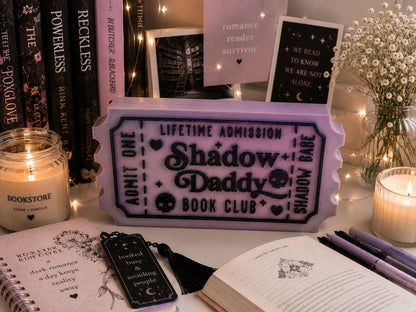 a purple ticket-shaped book club sign, surrounded by various items such as candles, books, and a vase with flowers.