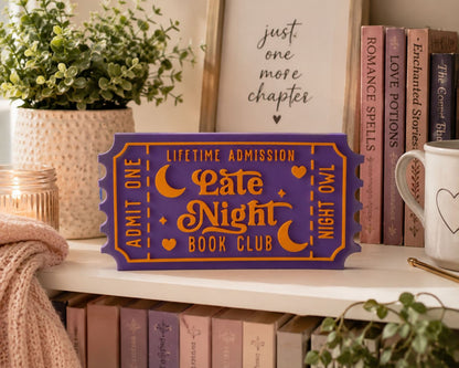 a purple ticket-shaped sign with orange and white text, placed on a shelf among books and a plant.