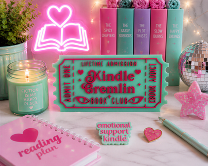 a pink Kindle e-reader, a pink notebook, a pink candle, a pink star, and a pink heart-shaped badge, all arranged on a marble surface.