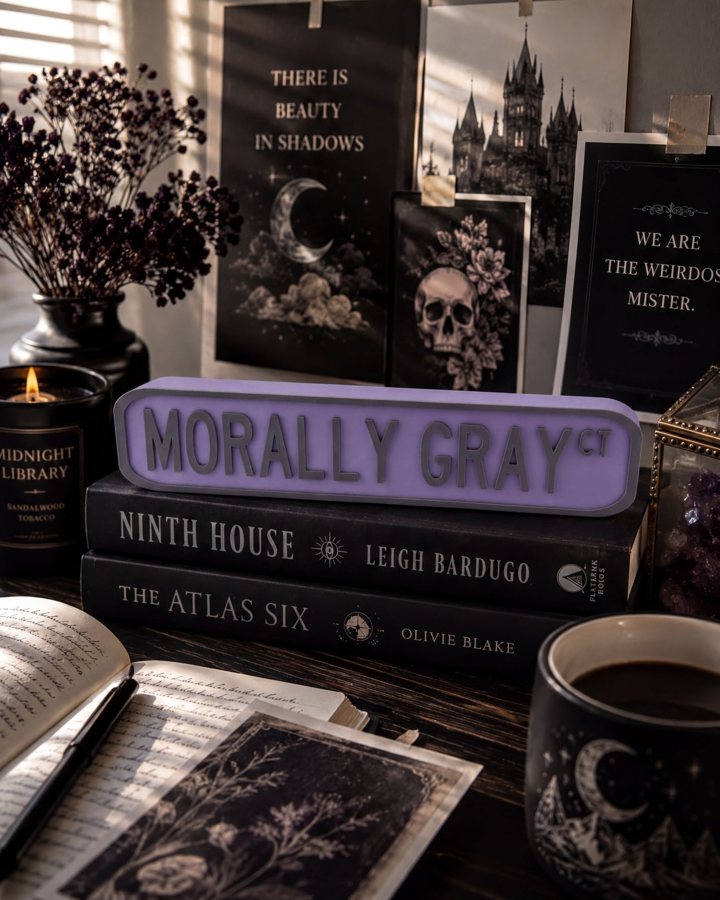 a desk with various items, including a stack of books, a mug, a candle, and a sign that says "Morally Gray". There is also a vase with flowers and a skull decoration on the desk.