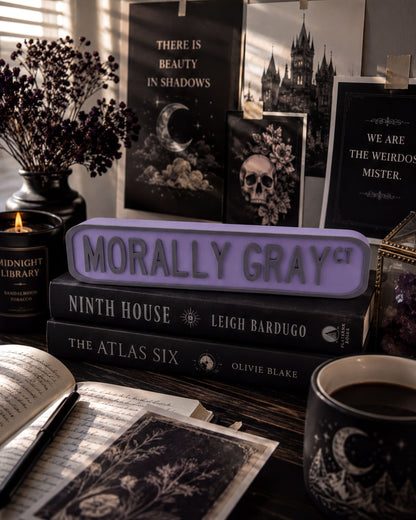 a desk with various items, including a stack of books, a mug, a candle, and a sign that says "Morally Gray". There is also a vase with flowers and a skull decoration on the desk.