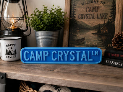 a wooden shelf with various items, including a blue sign that says "CAMP CRYSTAL", a lantern, a potted plant, and a framed picture of a cabin.