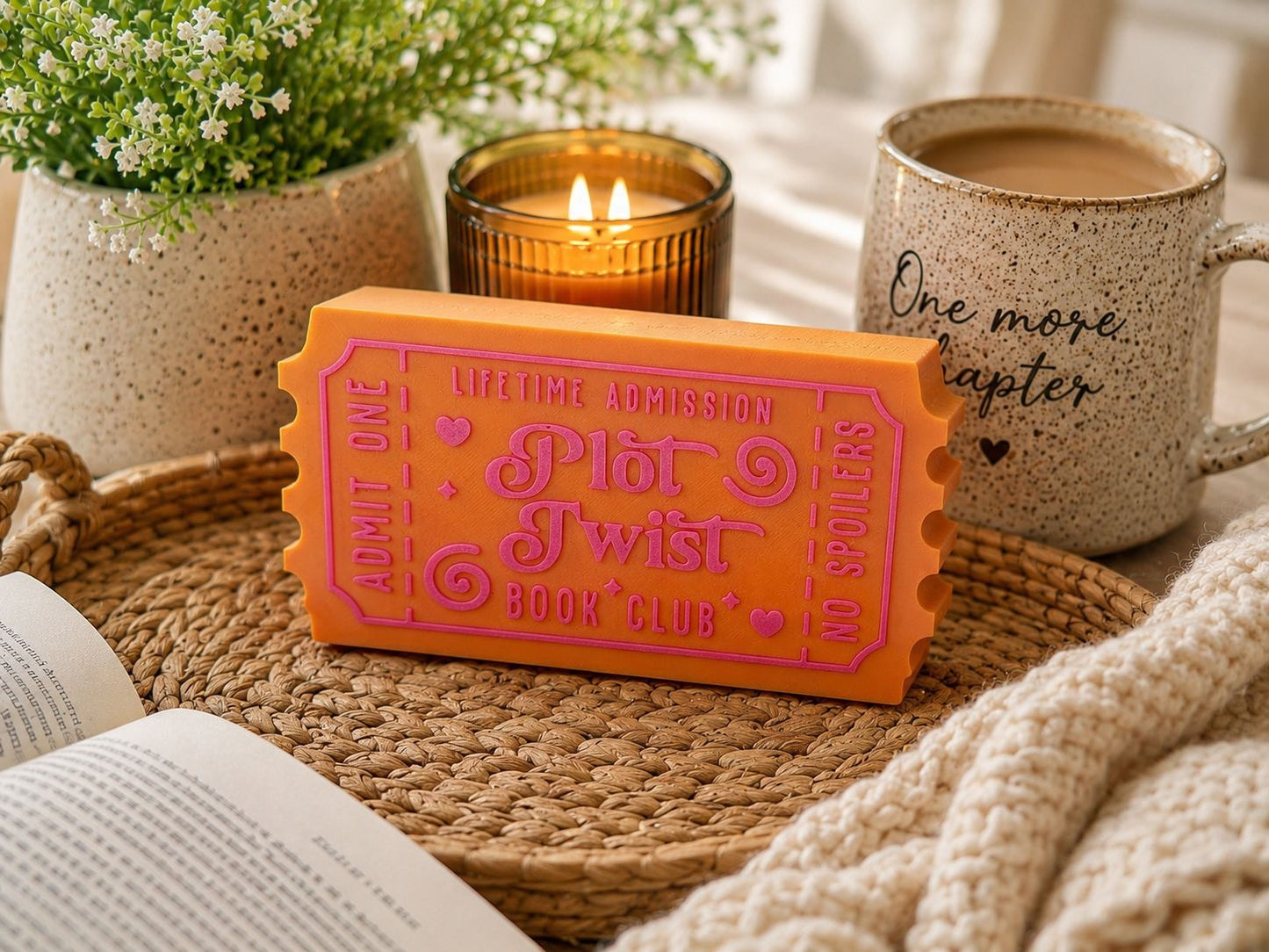 a cozy scene with a book, a mug, a candle, and a ticket-shaped box on a woven tray.
